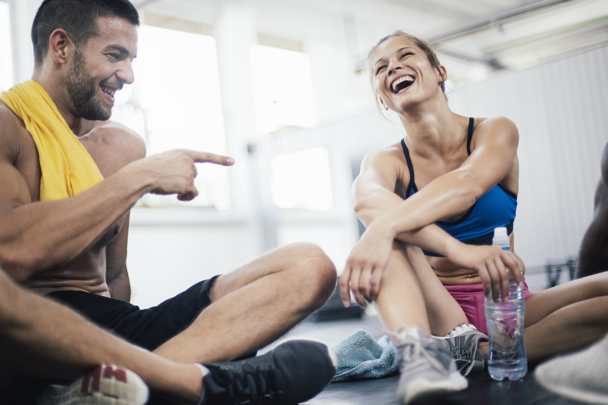 Ein Mann und eine Frau sitzen in Sportkleidung lachend auf dem Boden eines Fitnessstudios und entspannen sich nach dem Training.