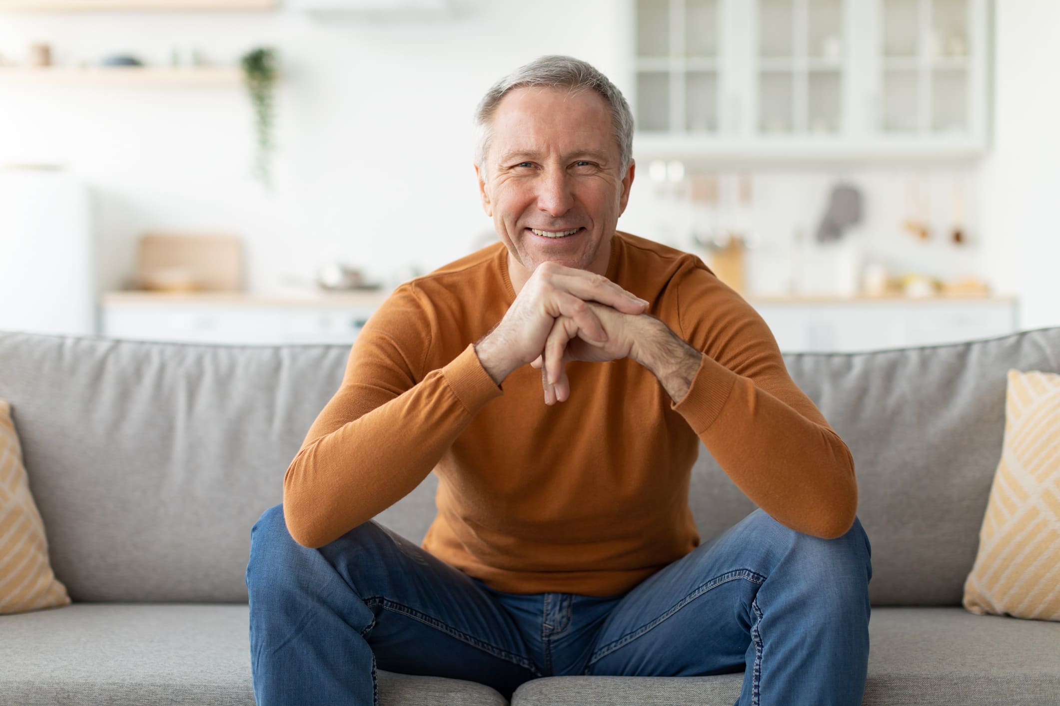 Ein älterer Mann sitzt entspannt auf einem Sofa, lächelt selbstbewusst und strahlt Zufriedenheit aus.